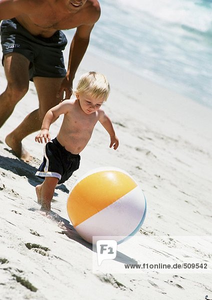 Man following child kicking ball at the beach.