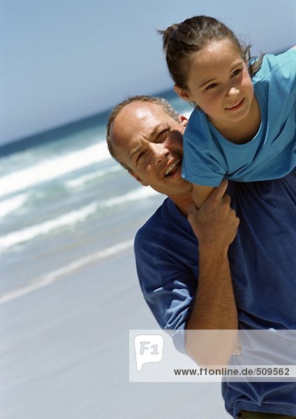 Mann mit jungem Mädchen auf der Schulter am Strand.