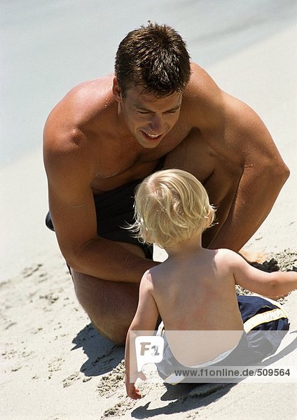 Rear view of young child sitting on beach looking at man.