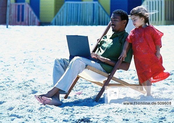 Mann sitzt auf einem Stuhl am Strand mit Laptop  kleines Mädchen steht an seiner Seite