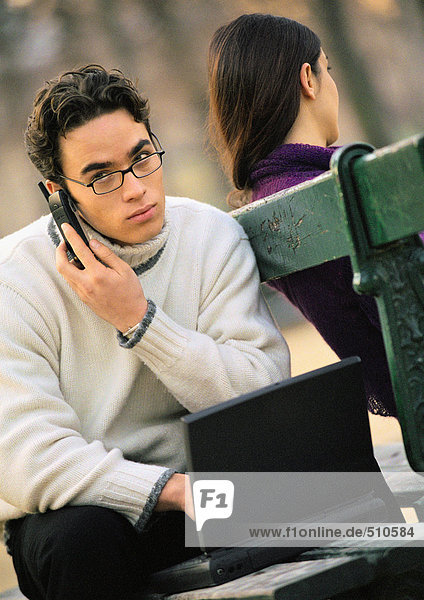 Man sitting on park bench with cell phone and laptop computer  woman sitting on other side of bench