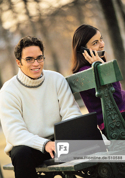 Man sitting on bench with laptop computer and woman using cell phone