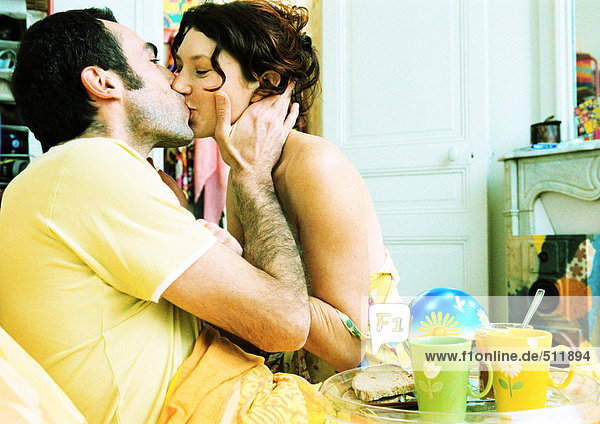 Mann im Bett mit Frühstückstablett auf dem Schoß  küssende Frau  Seitenansicht.