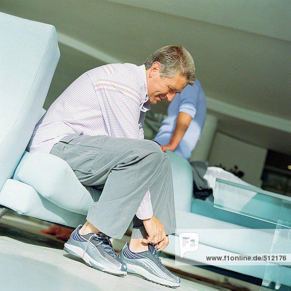 Erwachsener Mann  der Schuhe anzieht  zweite Person  die sich auf den Arm des Sofas lehnt  Seitenansicht