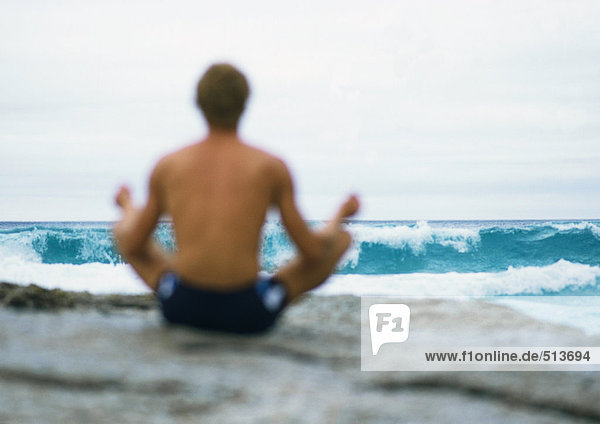 Mann sitzt in Lotusstellung vor dem Meer  Rückansicht  Fokus auf Welle im Hintergrund