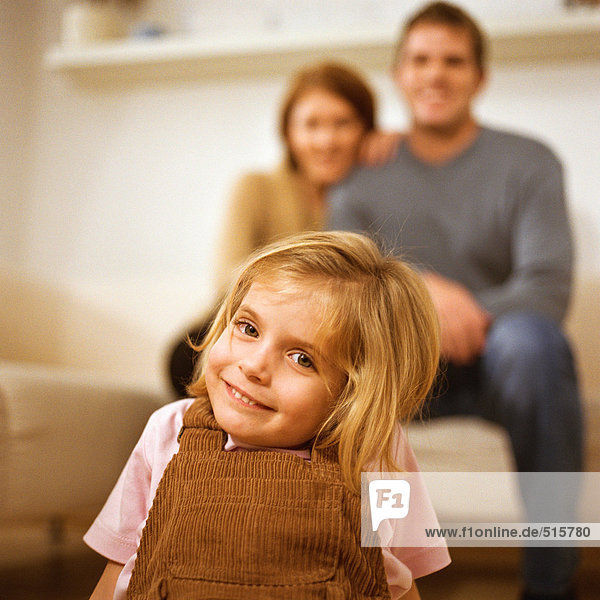 Kleines Mädchen lächelt vor der Kamera  Eltern hinter ihr