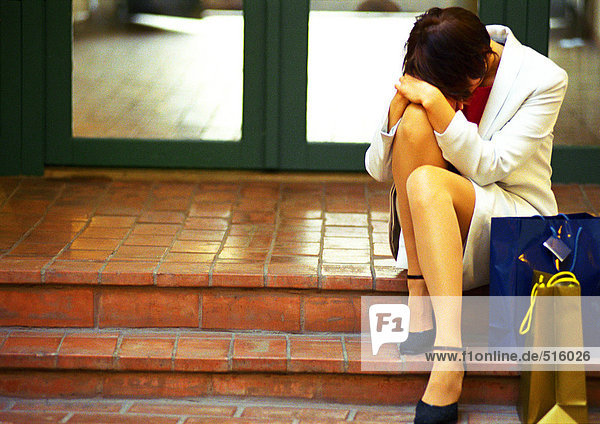 Woman in business clothes sitting on steps with head down  hands on knee  bags to the side.