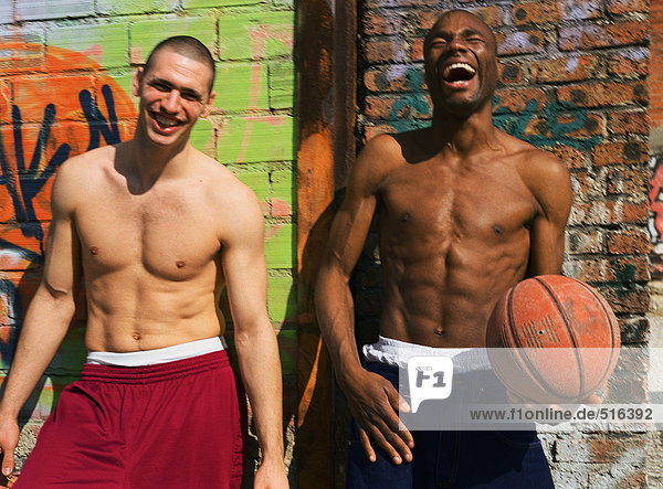 Zwei Männer ohne Hemd stehen und lachen neben der graffitierten Wand  einer hält Basketball.