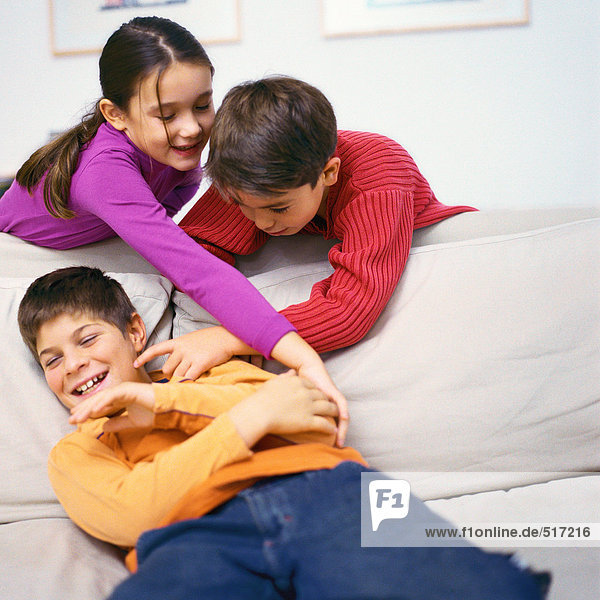 Junge und Mädchen lehnen sich über das Sofa und kitzeln den zweiten Jungen auf dem Sofa.
