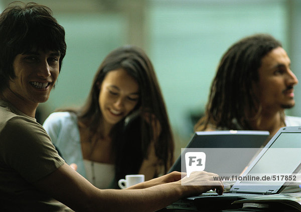 Man working on laptop  smiling at camera  with woman on phone and second man looking away