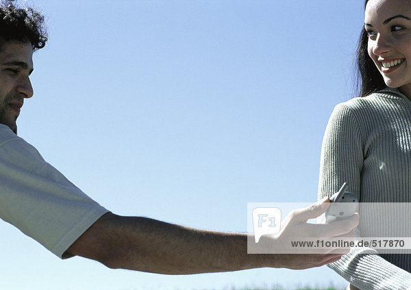 Junger Mann übergibt Handy an junge Frau mit Himmel im Hintergrund