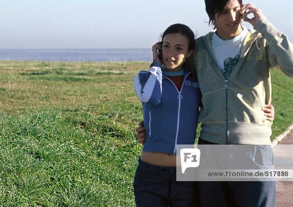 Young couple standing with arms around eachother  both talking on cell phones