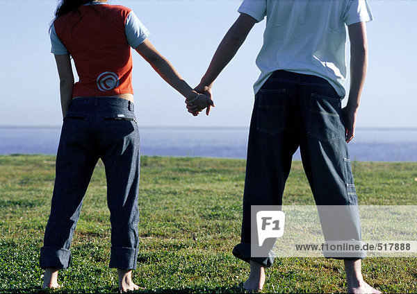 Junges Paar hält sich an den Händen  steht Seite an Seite und betrachtet das Meer.