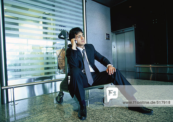 Businessman sitting on luggage cart talking on cell phone.