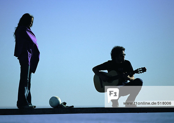 Frau stehend  Mann hockend  Gitarre auf Bank spielend