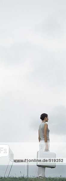 Frau auf Gras stehend  hält metallische Aktentasche vor dem Stuhl  Himmel im Hintergrund