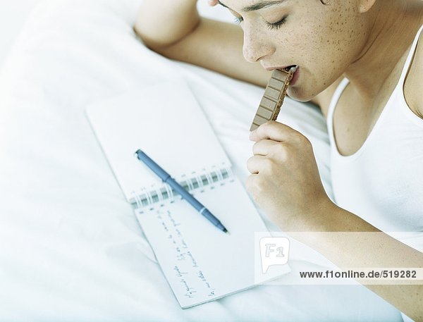 Frau liegt auf dem Bett  isst Schokolade und schaut auf das Notizbuch mit dem Stift darüber.