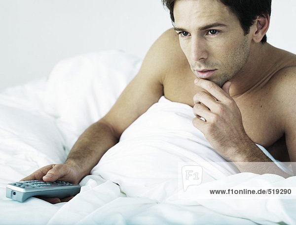 Man in bed under covers  sitting up  pointing remote control  touching chin