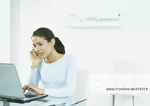 Woman sitting at table typing on laptop  holding cell phone to ear