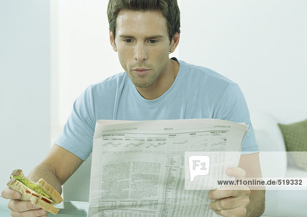 Young man reading newspaper and eating sandwich