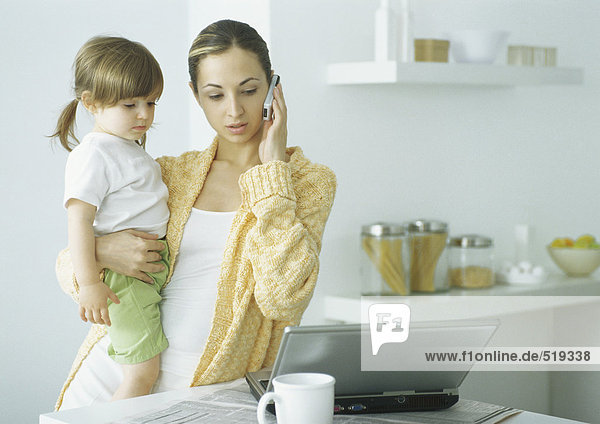 Junge Frau hält kleines Mädchen mit einem Arm  hält Handy an Ohr mit der anderen Hand  schaut auf Laptop