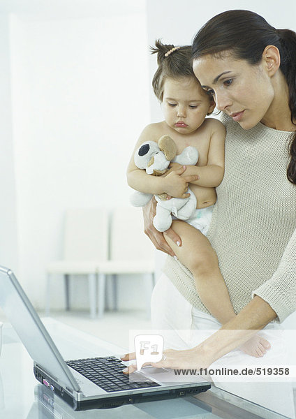 Frau hält kleines Mädchen in einem Arm,  Tippen auf dem Laptop mit der anderen Hand
