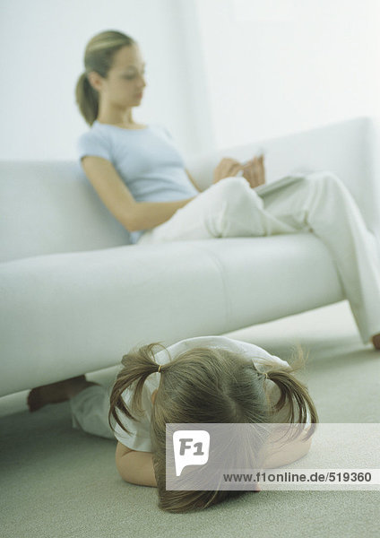 Kleines Mädchen auf dem Boden liegend,  junge Frau auf dem Sofa sitzend,  lesend