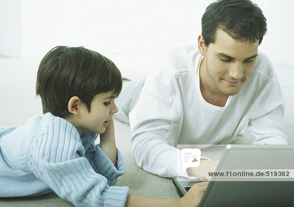 Man and little boy lying on floor on stomachs  working on laptop