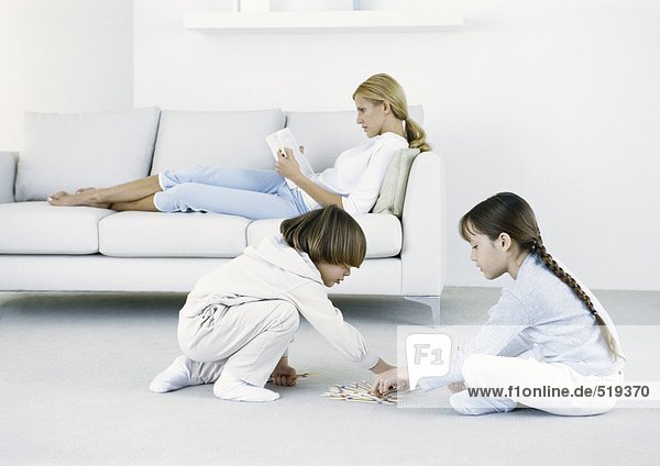 Junge und Mädchen sitzen auf dem Boden  Frau sitzt auf dem Sofa und liest im Hintergrund.