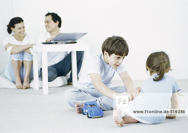 Junge und Mädchen spielen auf dem Boden  Eltern nutzen Laptop im Hintergrund