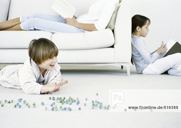 Junge spielt Murmeln auf dem Boden,  Frau und Mädchen lesen im Hintergrund