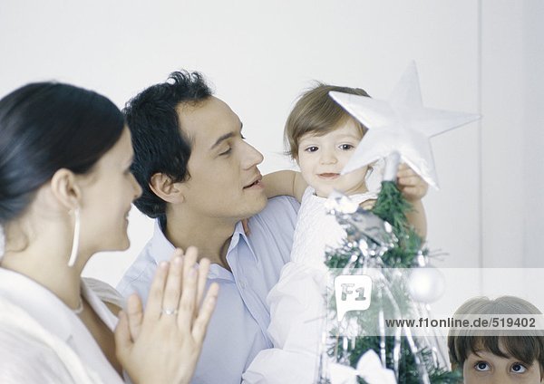 Kleines Mädchen in Vaters Armen berührt Stern am Weihnachtsbaum neben Mutter und Bruder