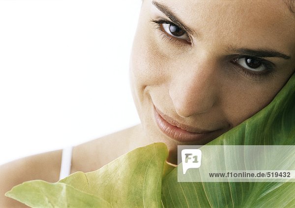 Frau kippt Kopf gegen grünes Blatt  lächelt in die Kamera  Nahaufnahme