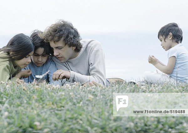 Mann auf Gras liegend mit Junge und Mädchen auf Handy schauend  zweiter Junge sitzend getrennt