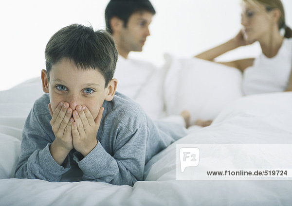 Junge auf dem Bauch liegend im Bett  Mund mit Händen bedeckend  Eltern diskutieren im Hintergrund
