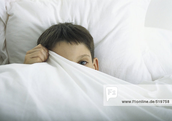 Boy in bed  peeking out behind sheet with one eye