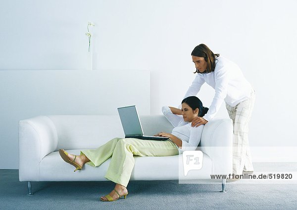 Woman lounging on sofa and using laptop  man standing  looking over woman's shoulder
