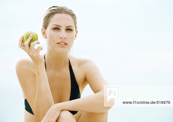 Junge Frau im Bikini sitzend mit hochgezogenen Knien und haltendem Apfel