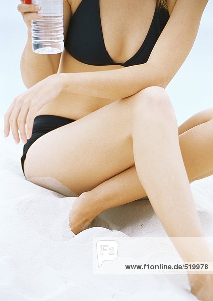 Junge Frau im Bikini sitzend im Sand  Wasserflasche haltend  Teilansicht