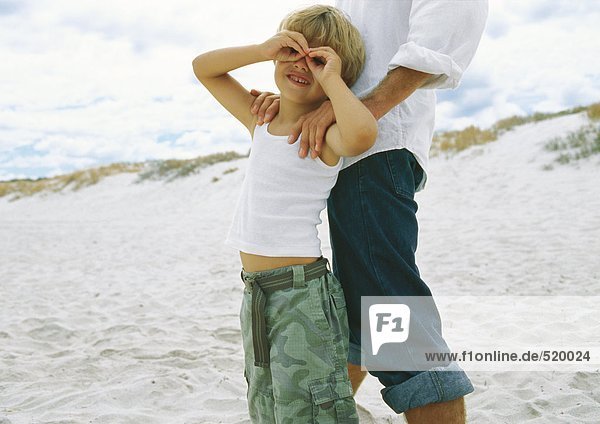 Junge hält Hände um die Augen  lehnt sich an Vater am Strand an