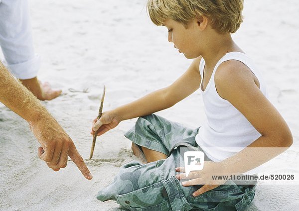 Junge auf Sand sitzend  Zeichnung mit Stock  Vater zeigend
