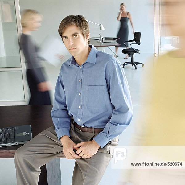 Man sitting on edge of desk in busy office