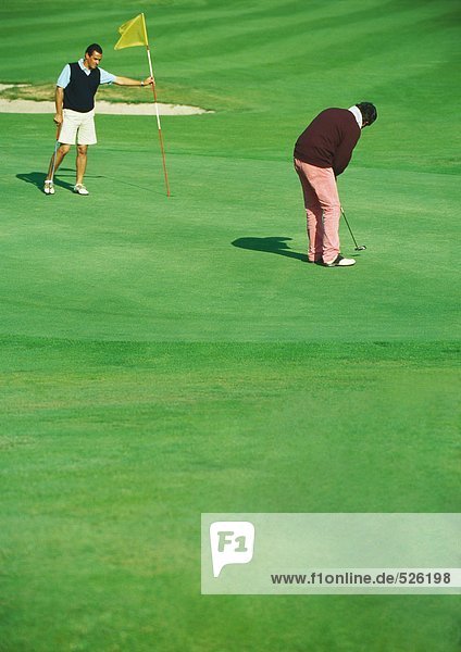 Golfer putting while second man holds golf flag