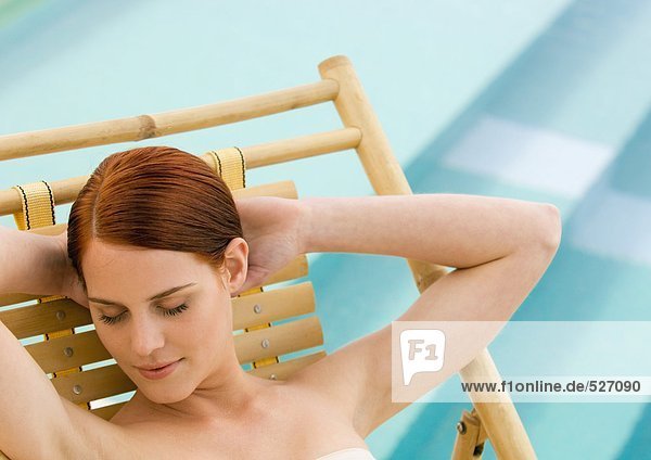 Frau im Liegestuhl sitzend,  Hände hinter dem Kopf und Augen geschlossen,  Pool im Hintergrund