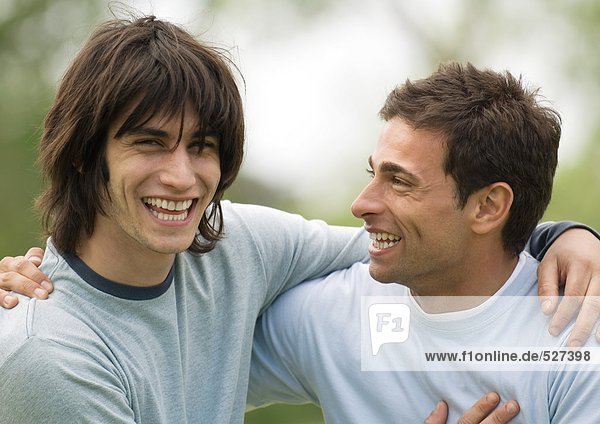 Zwei junge männliche Freunde lächeln mit den Armen um die Schultern.