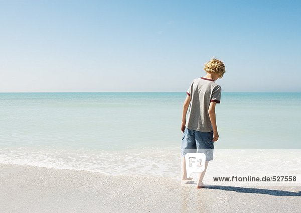 Junge testet Wasser mit Zehe am Strand  Rückansicht