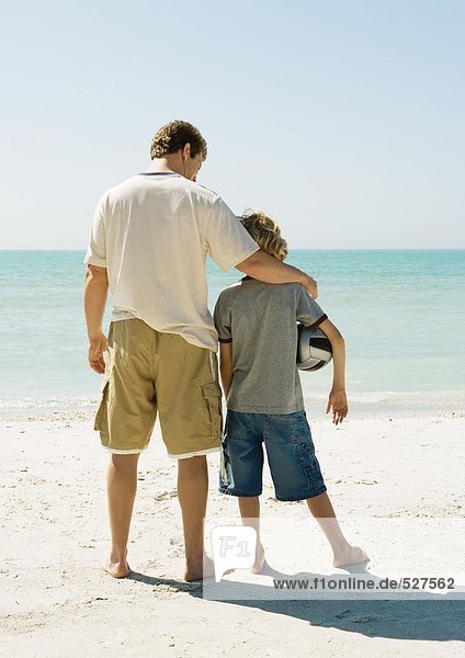 Vater und Sohn stehen zusammen am Strand  Junge hält Ball unter Arm