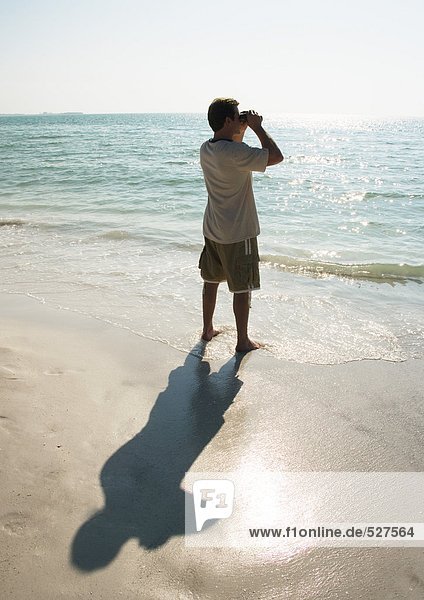 Mann steht am Strand und schaut mit einem Fernglas auf das Meer.