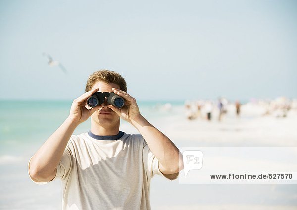 Mann schaut durchs Fernglas am Strand  mit Blick auf die Kamera