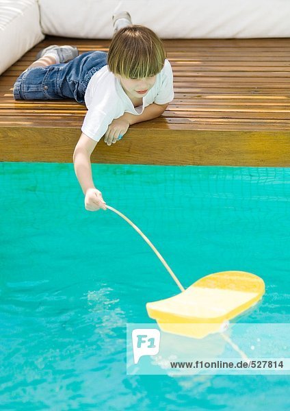 Junge liegt auf dem Boden in der Nähe des Pools und spielt mit einem Spielzeugboot.